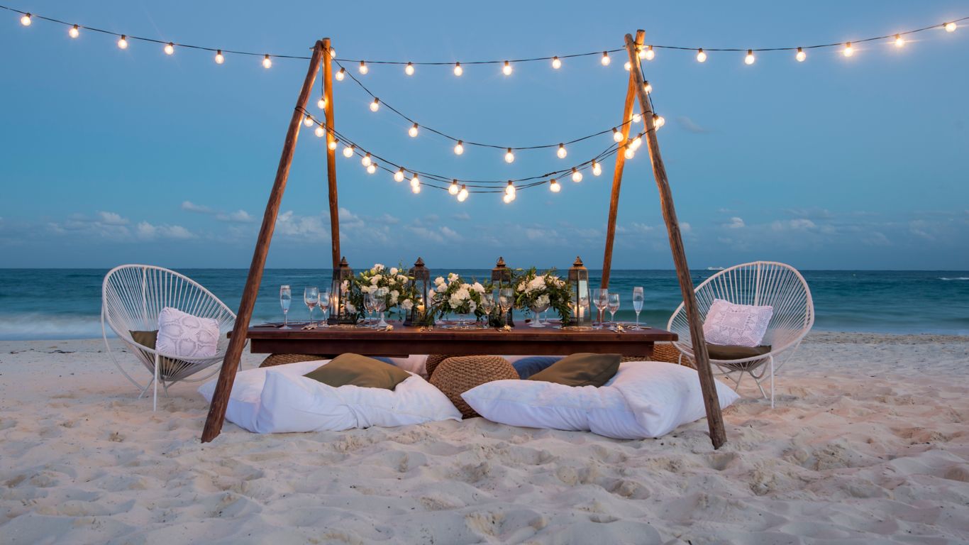 romantic dinner table for two arranged with flowers and string lights on the beach with the ocean in the backround